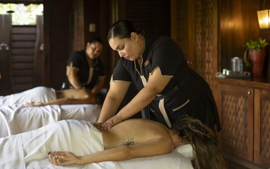Two massage therapists in a wooden spa room give massages to clients lying face down on tables, covered with white sheets, creating a relaxing and serene atmosphere.