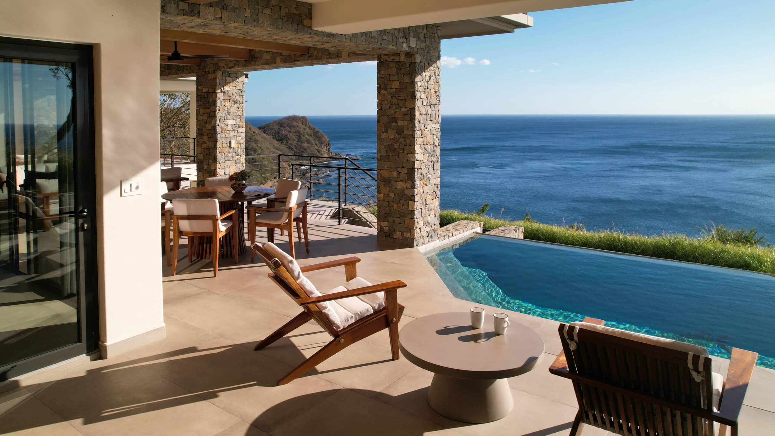 http://A%20modern%20patio%20with%20wooden%20chairs%20and%20tables%20overlooks%20an%20infinity%20pool%20and%20a%20vast%20blue%20ocean,%20with%20rocky%20cliffs%20and%20clear%20skies%20in%20the%20background.