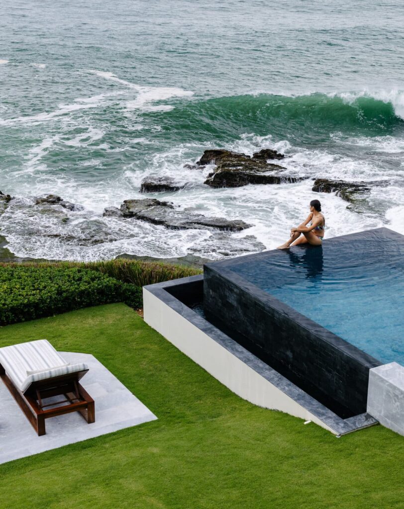 A person sits on the edge of an infinity pool overlooking the ocean, with waves crashing below. Two lounge chairs rest on nearby grass—perfect for unwinding and enjoying a special offer like a 3rd night free at this dreamy escape.