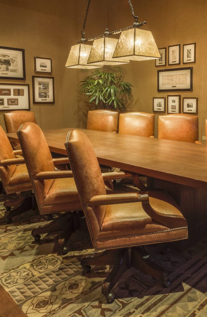 A conference room designed for group experiences, featuring a long wooden table surrounded by eight brown leather chairs, a patterned rug, framed pictures on the walls, a telescope in the corner, and a plant under warm lighting.