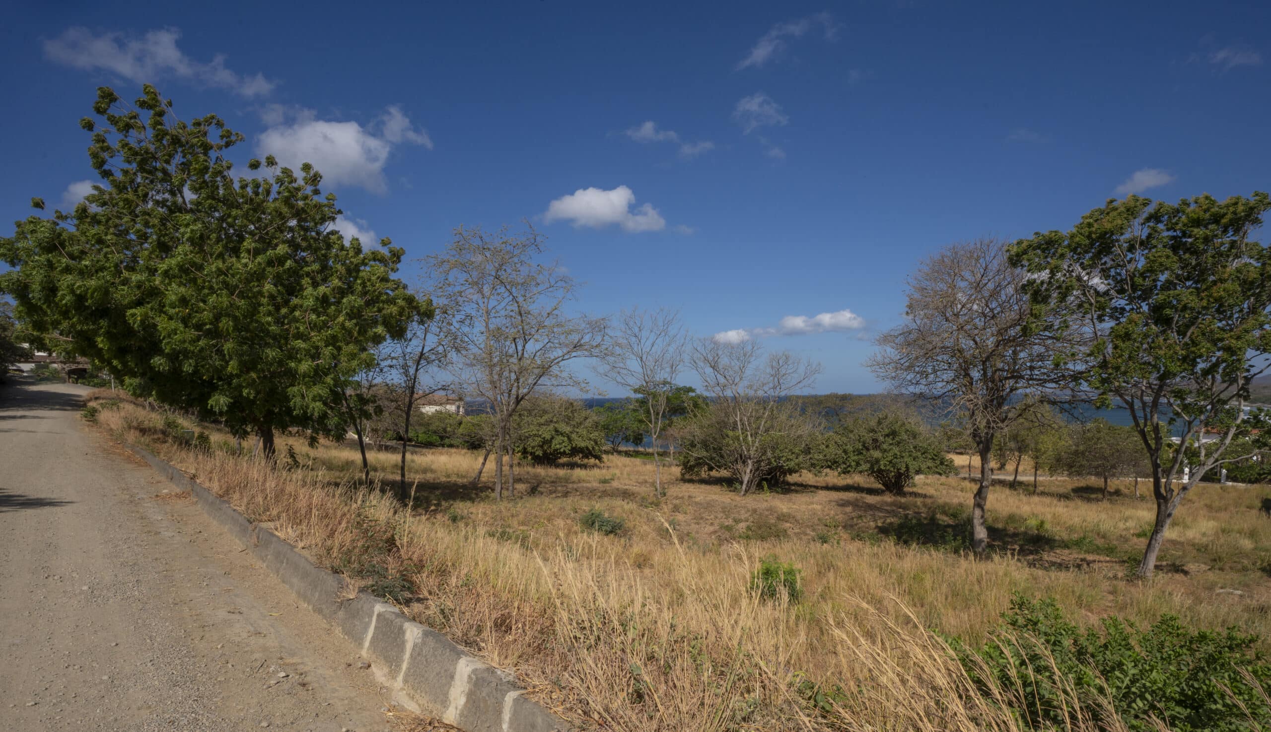 http://A%20dirt%20road%20runs%20alongside%20a%20dry,%20grassy%20field%20near%20Lot%20F9,%20with%20scattered%20trees%20beneath%20a%20clear%20blue%20sky.%20In%20the%20distance,%20glimmers%20of%20water%20and%20greenery%20enhance%20the%20serene%20landscape.