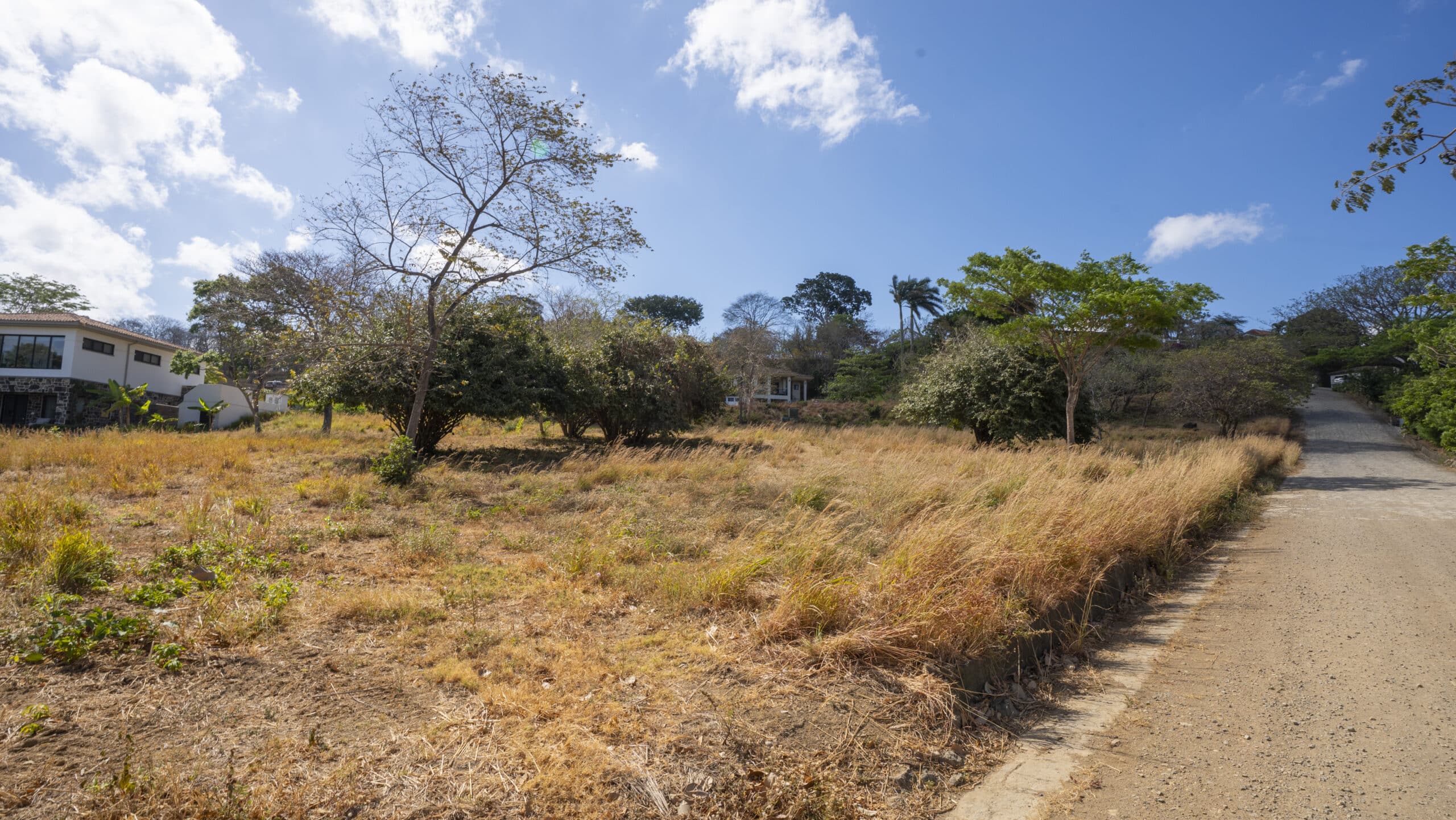 http://A%20dirt%20road%20runs%20beside%20a%20dry,%20grassy%20field%20with%20scattered%20trees%20and%20bushes.%20Modern%20houses,%20including%20those%20on%20Lot%20F8%20and%20Lot%20F9,%20are%20partially%20visible%20among%20the%20greenery%20beneath%20a%20bright%20blue%20sky%20with%20white%20clouds.
