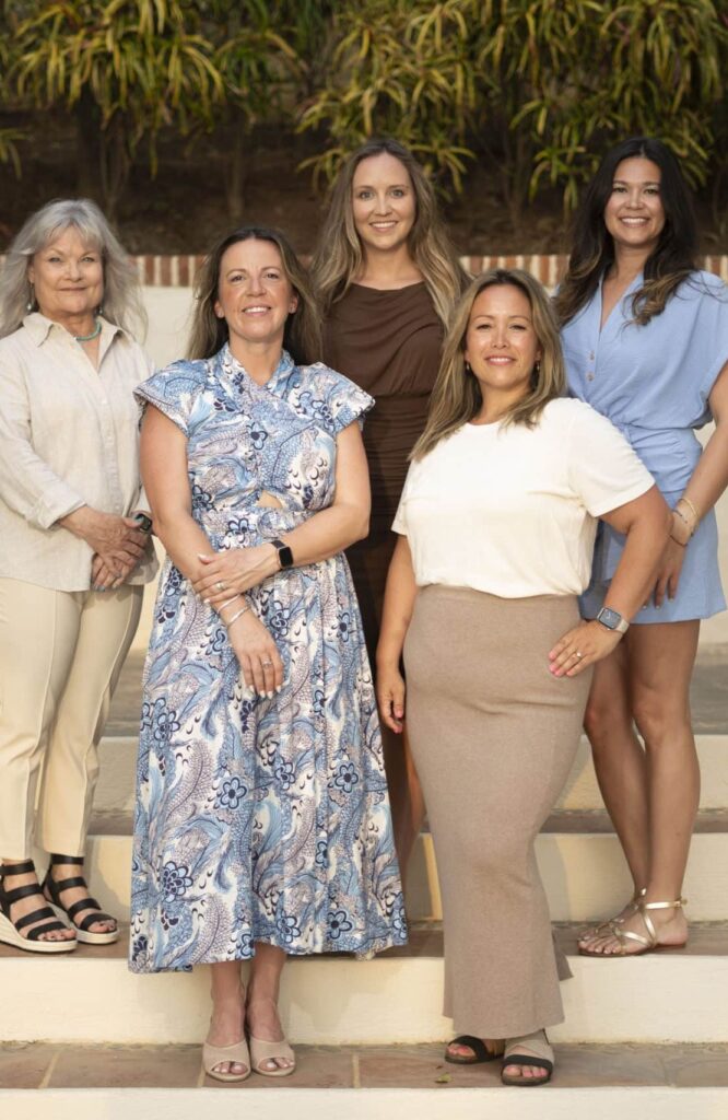 Five women stand together on outdoor steps, smiling at the camera. Dressed in light, summery clothing, they bring your team spirit to the Emerald Coast, with palm fronds and warm sunlight brightening the vibrant scene.