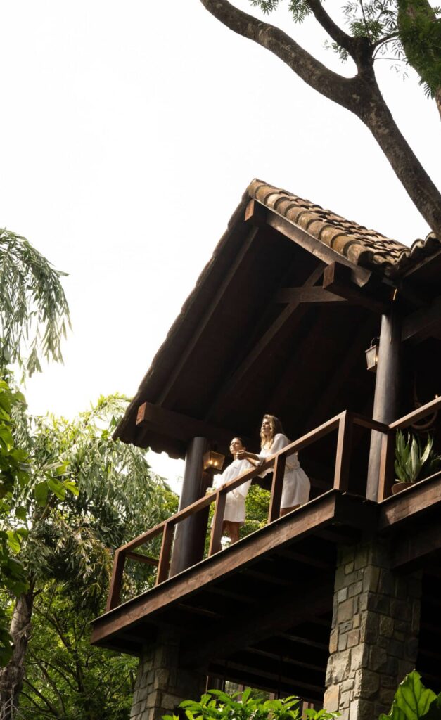 Two people stand on the balcony of a rustic stone and wood house, surrounded by lush green trees, enjoying the scenery on a bright day—an ideal escape for those seeking relaxing vacation packages.