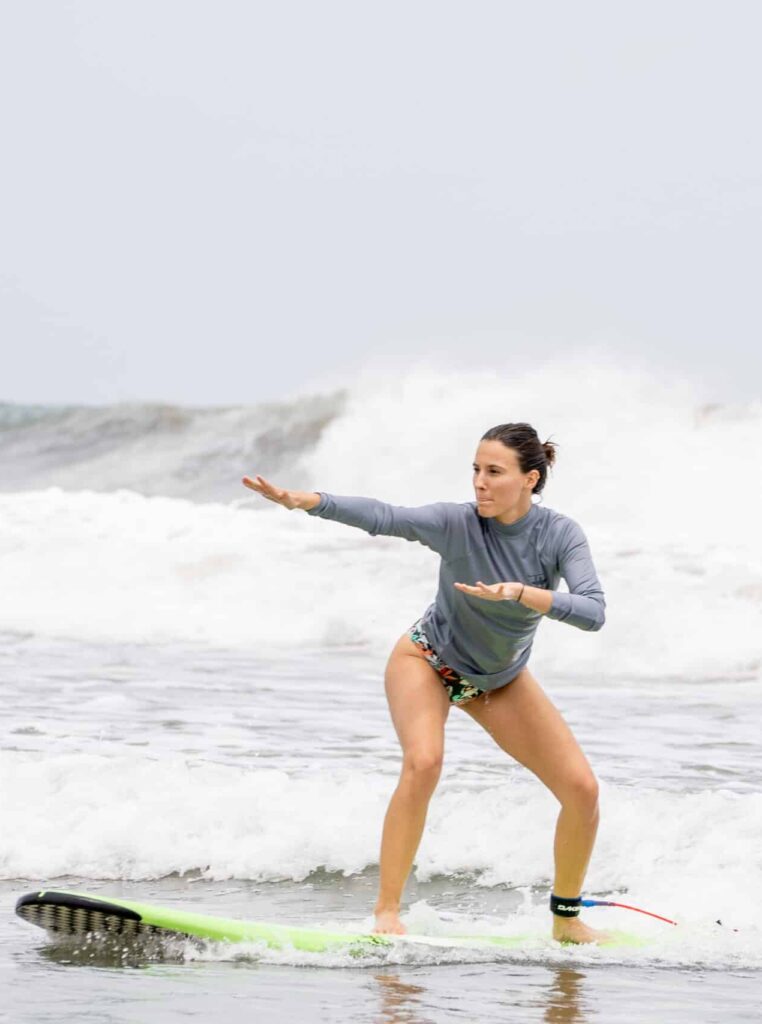 A woman in a gray long-sleeve top and patterned shorts balances on a green surfboard, riding a small wave in the ocean—this scene captures the adventure awaiting with our exclusive vacation packages.