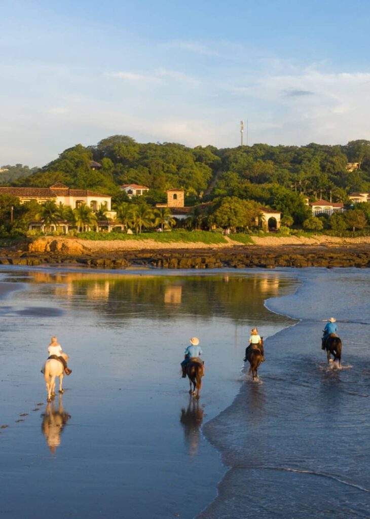 Five people ride horses along a sandy beach at sunset, with lush green hills and houses in the background. The calm waves and clear sky create a tranquil atmosphere—perfect for unforgettable vacation packages.