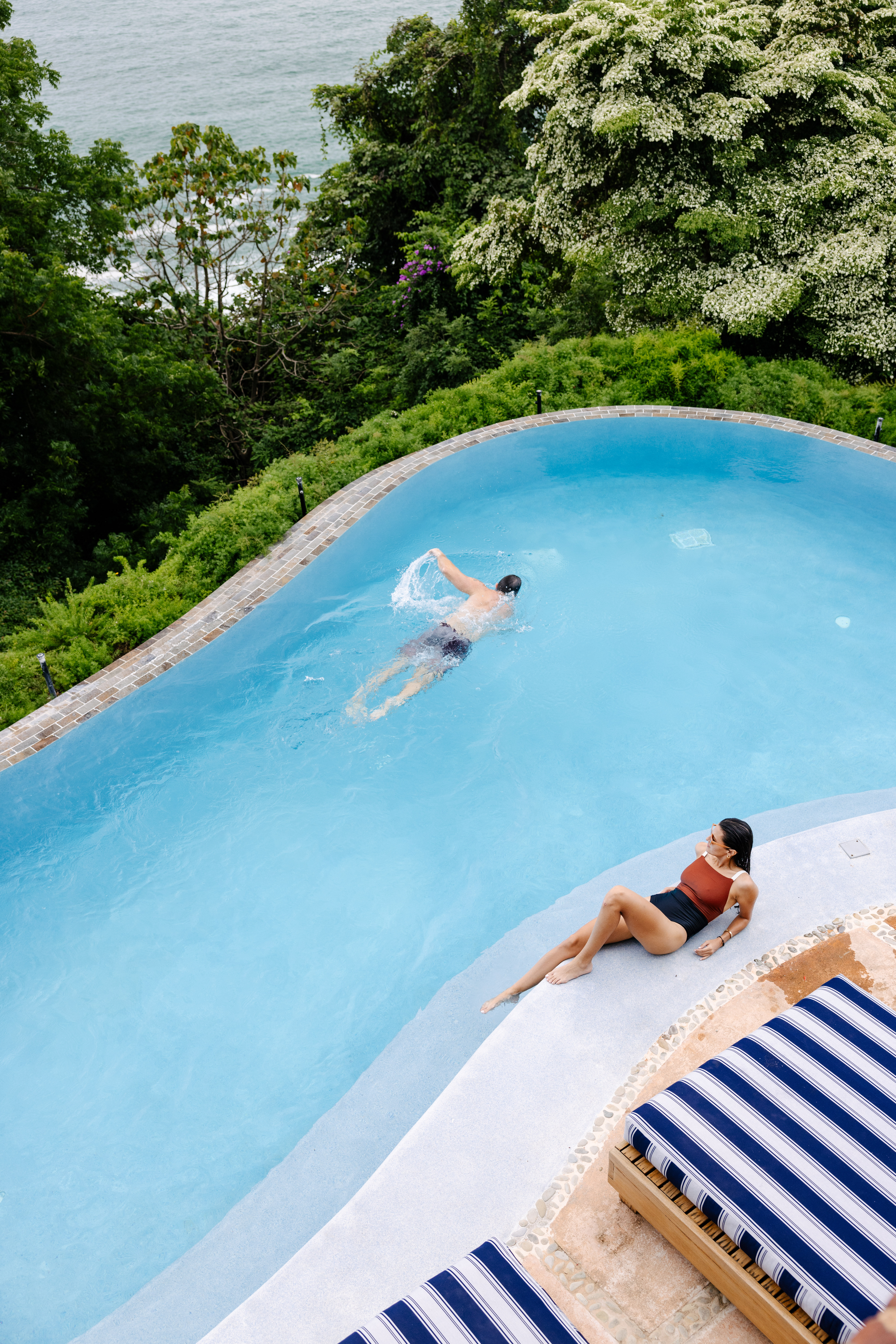 A person swims in a curved outdoor pool while another relaxes poolside in a swimsuit. Blue striped lounge chairs invite you to stay longer, surrounded by lush greenery and water views—perfect for enjoying your 5th night free.