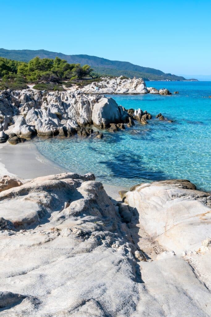 Rocky coastline with clear turquoise water, sandy beach, and lush green hills under a bright blue sky make this hidden vacation spot perfect for those seeking tranquility away from the crowds.