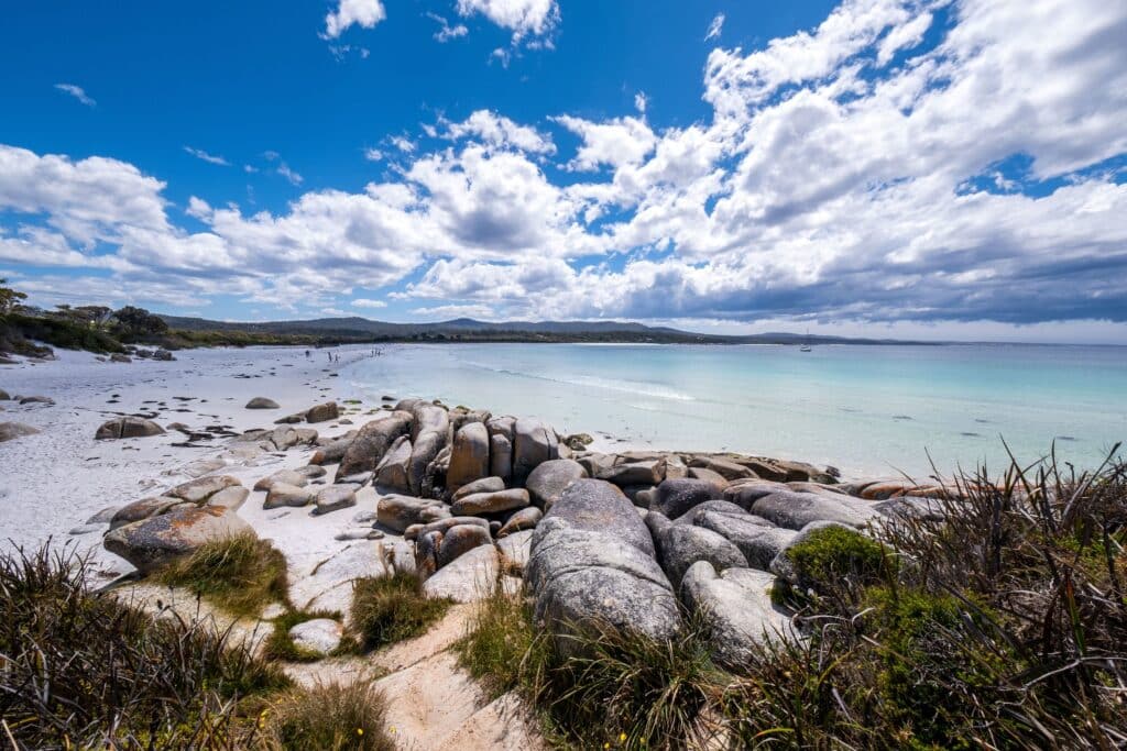 A scenic beach view with large smooth rocks on white sand, clear turquoise water, and a partly cloudy blue sky. Sparse grass grows near the rocks along this coastline—one of the region’s true under-the-radar destinations.