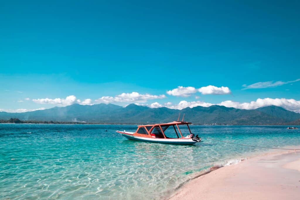A small boat with a red roof floats on clear turquoise water near a sandy beach, making it one of those under-the-radar destinations perfect for luxury travel, with distant mountains and a bright blue sky in the background.