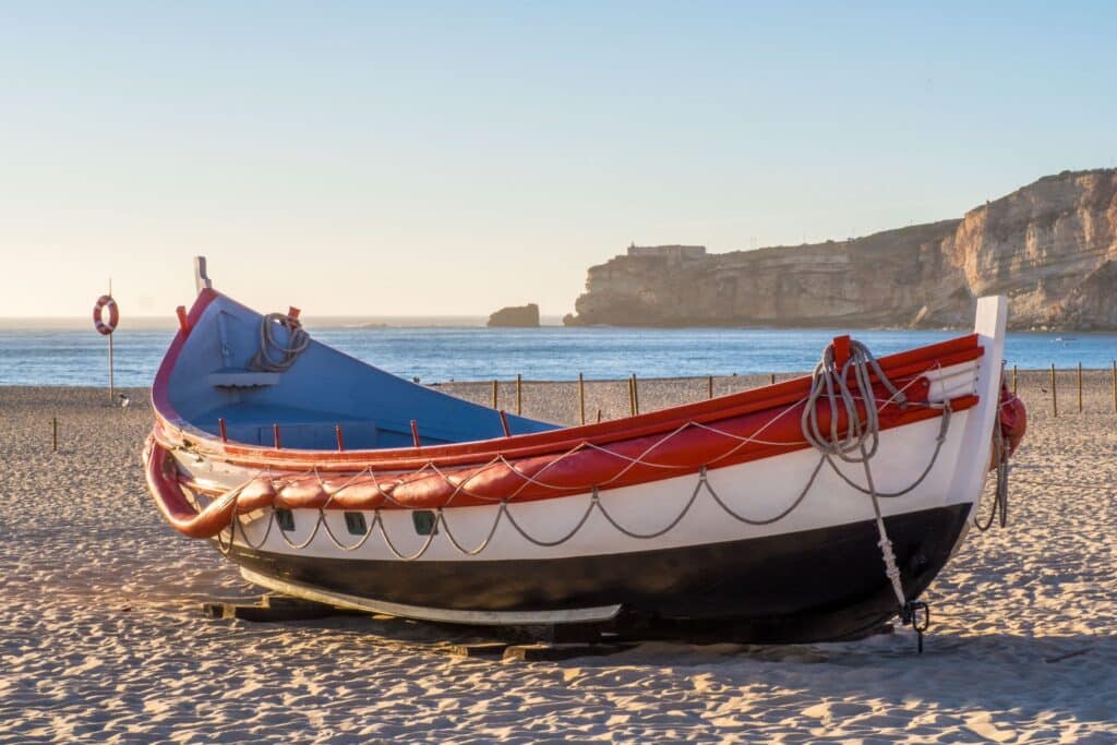 A colorful wooden boat with red, white, and blue paint sits on a sandy beach in sunlight—an inviting scene for those seeking under-the-radar travel destinations. The sea and rocky cliffs are visible in the background under a clear sky.