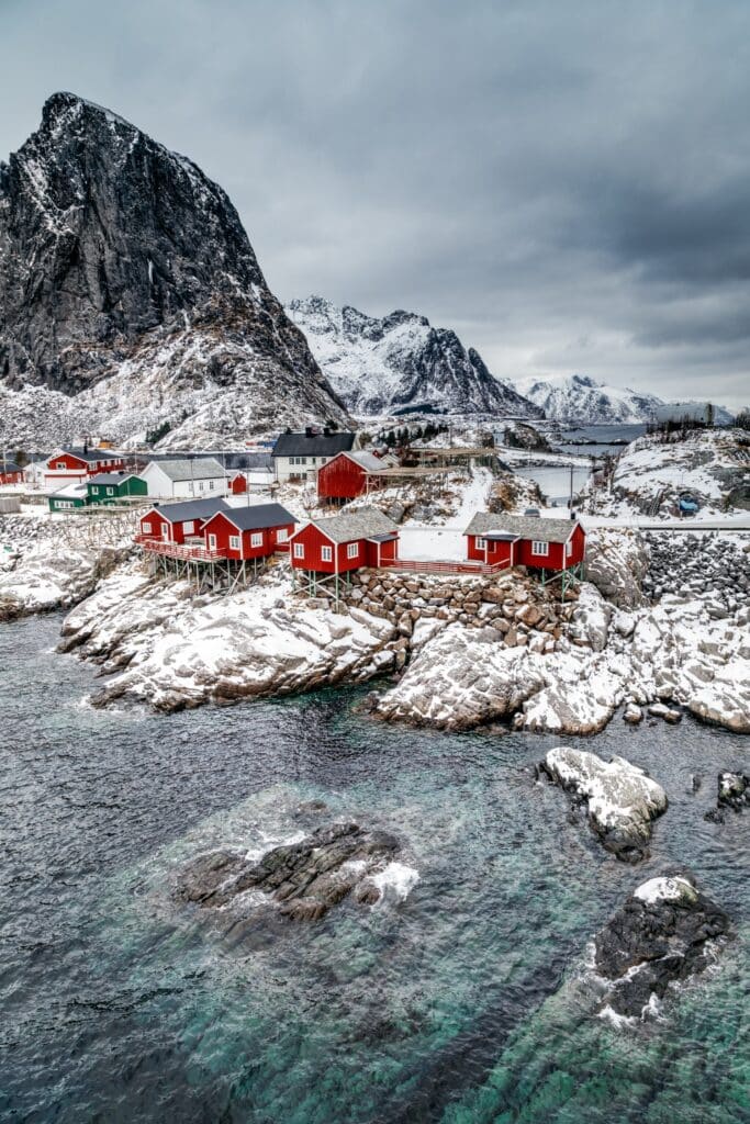 Red wooden cabins sit on a rocky, snow-dusted shoreline by clear turquoise waters, with tall mountains and dramatic cloudy skies in the background, capturing an under-the-radar travel destination for a wintery coastal village escape.