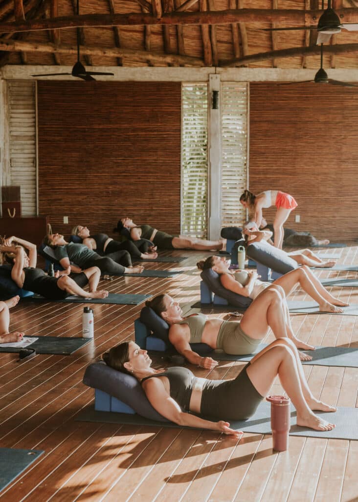 A group enjoys a restorative yoga session on mats with bolsters under their backs in a sunlit wooden studio. Highlighting group experiences, an instructor assists one participant, while water bottles rest beside some mats.
