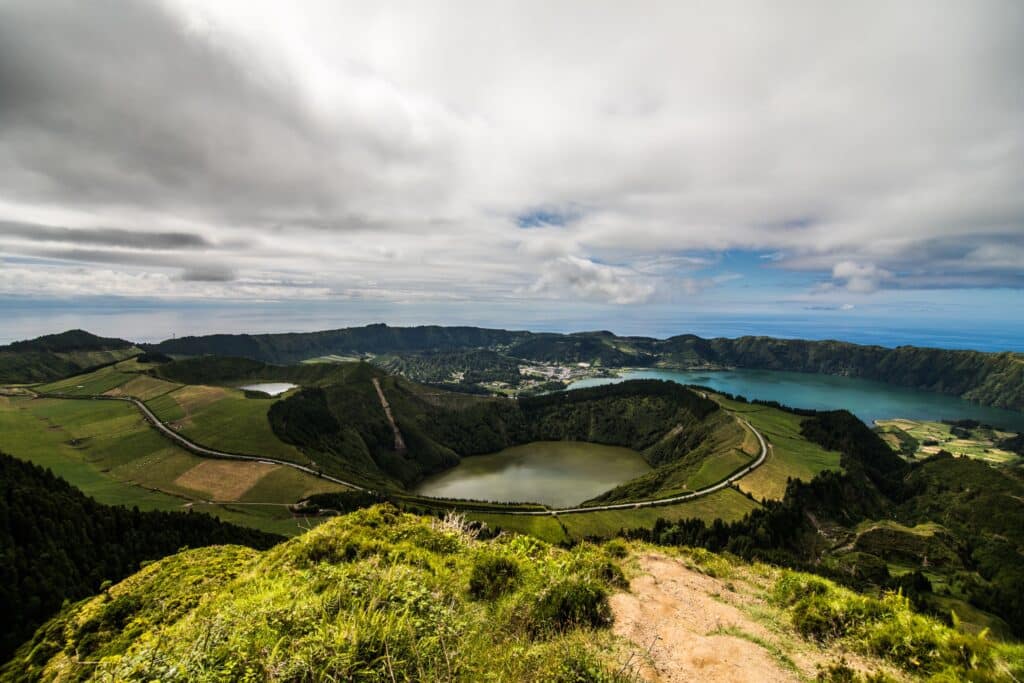 A scenic view from a high vantage point shows lush green hills, two crater lakes of different colors, and a small village nestled between them—an under-the-radar destination perfect for unique travel experiences under a partly cloudy sky.