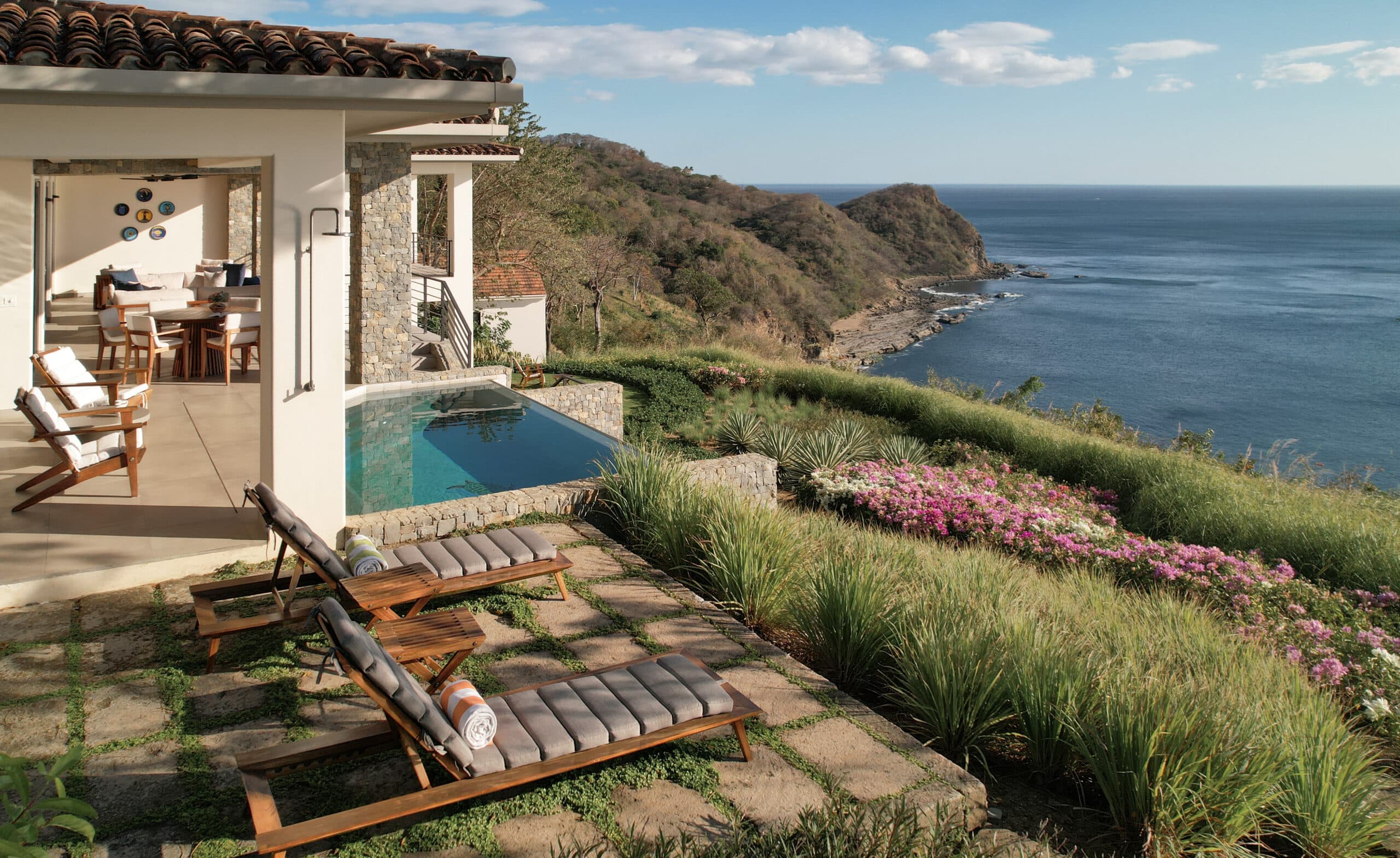 A luxurious co-owned luxury home, Casa Tucan features an infinity pool overlooking the ocean and rocky coastline. Two cushioned lounge chairs rest on a stone patio amid lush grass and flowers under a clear sky.