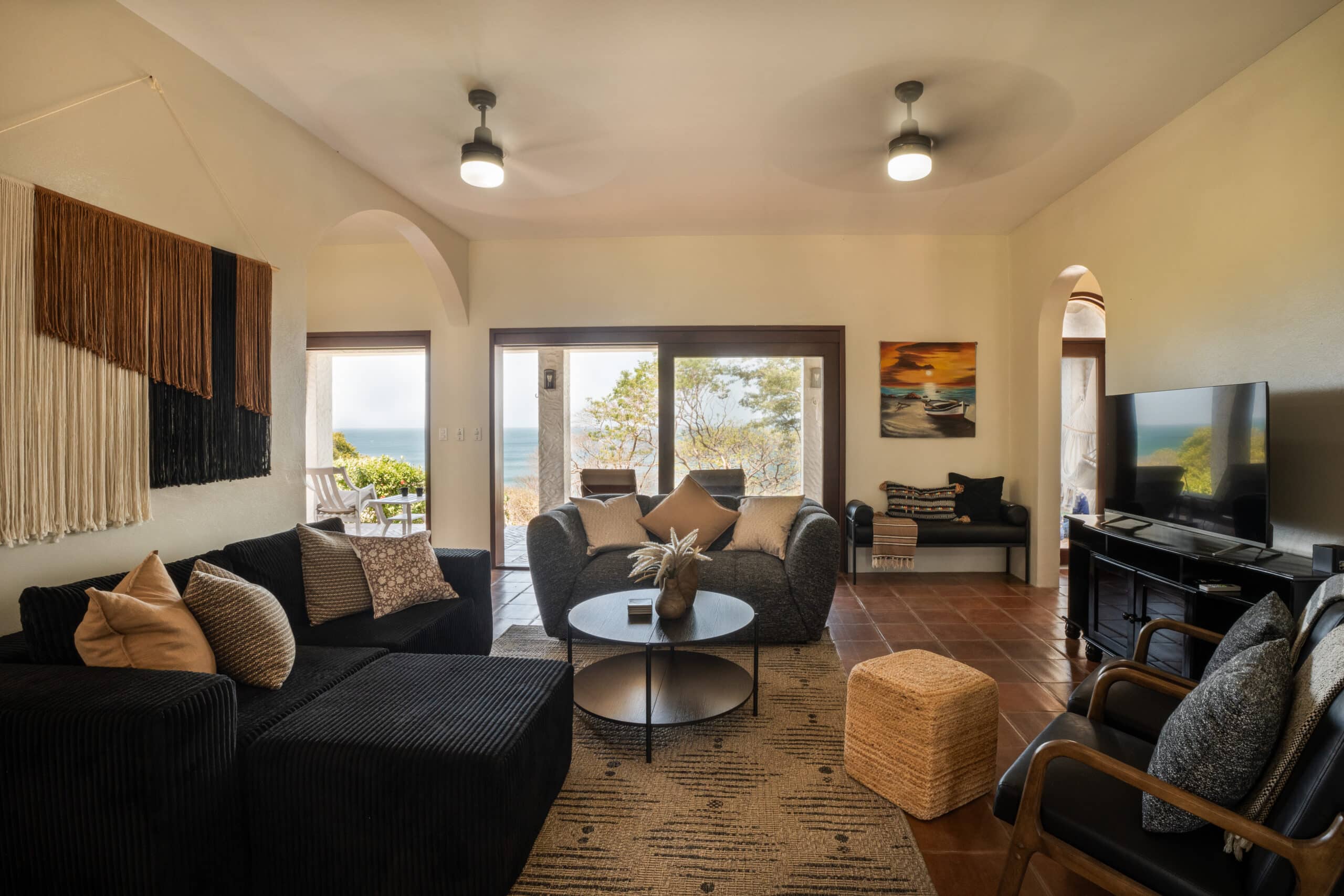 http://A%20cozy%20living%20room%20at%20Rosada%20Seascape%20features%20black%20sofas,%20patterned%20cushions,%20a%20round%20coffee%20table,%20and%20a%20TV.%20Large%20windows%20reveal%20the%20patio%20and%20ocean%20view,%20while%20warm%20lighting%20and%20modern%20decor%20create%20a%20welcoming%20atmosphere.