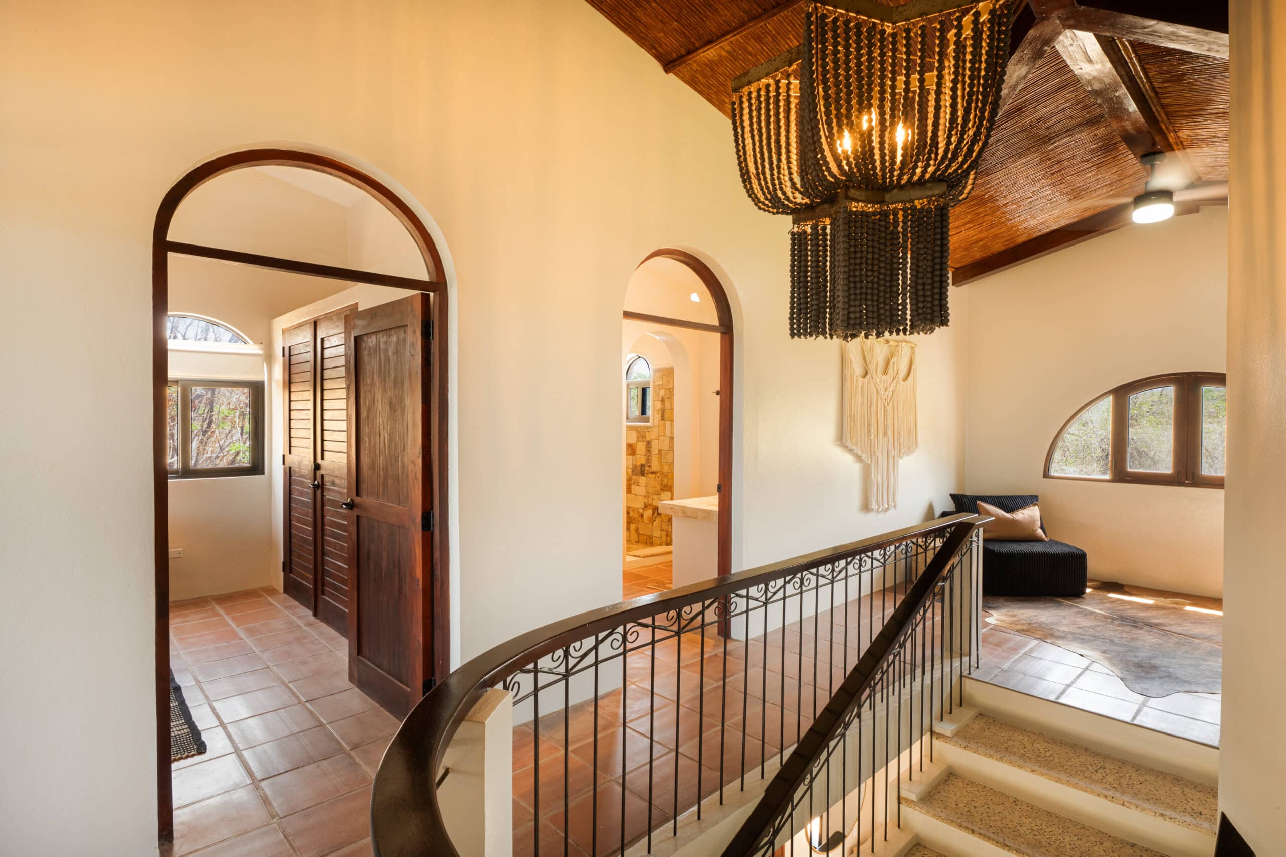http://A%20warmly%20lit%20hallway%20with%20arched%20doorways,%20a%20tiled%20floor,%20a%20black%20bead%20chandelier,%20B-25%20stair%20railing,%20wood%20ceiling,%20and%20a%20cozy%20sitting%20nook%20with%20cushions%20by%20an%20arched%20window.
