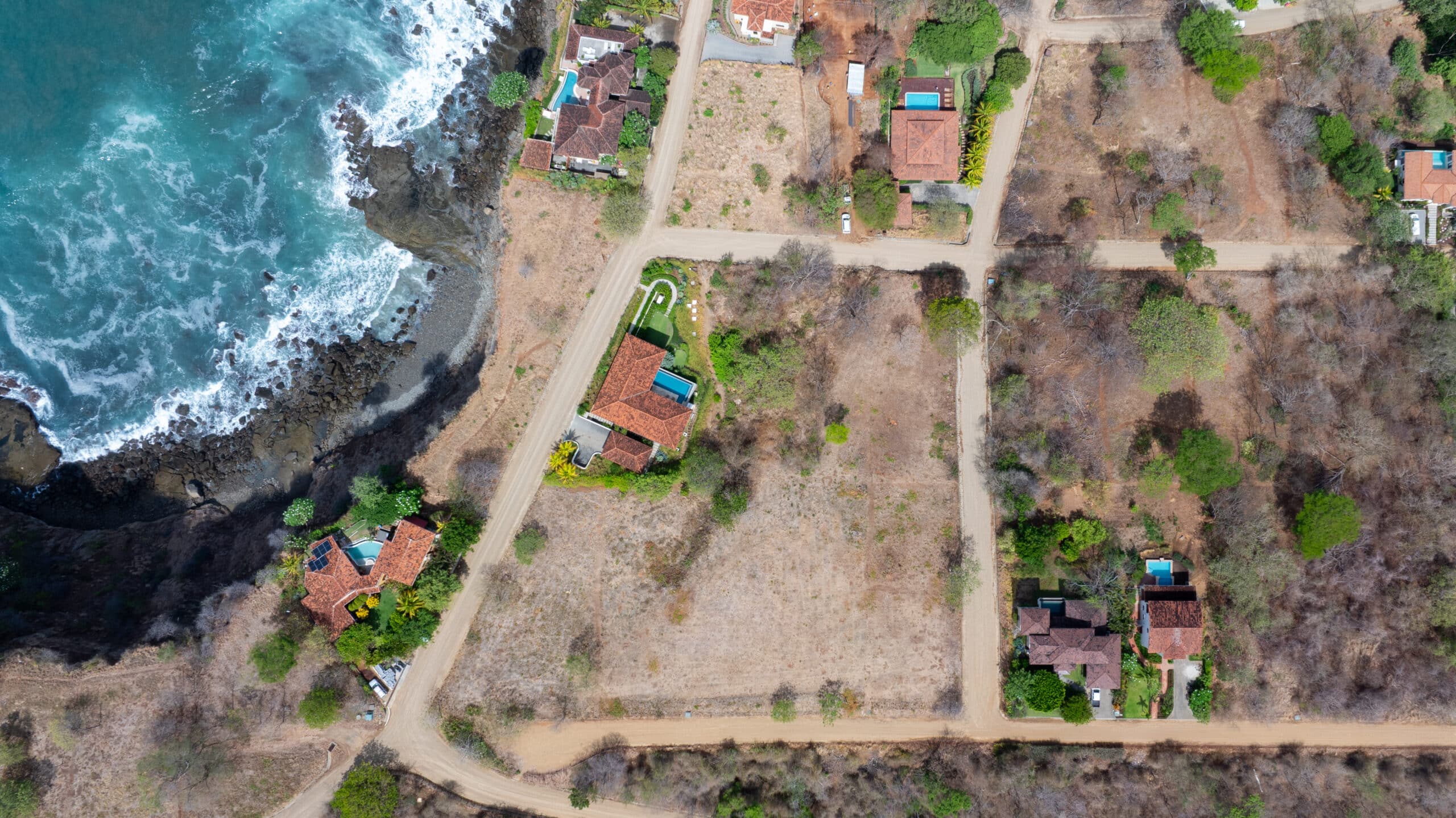 http://Aerial%20view%20of%20coastal%20homes%20with%20red%20tile%20roofs%20and%20blue%20swimming%20pools,%20nestled%20among%20dry%20brush%20and%20trees,%20with%20crashing%20waves%20on%20the%20rocky%20shore.