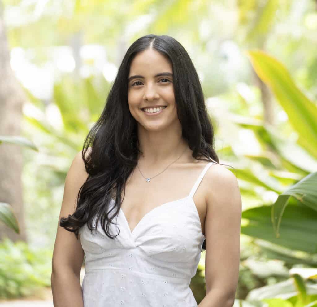 A woman with long dark hair, Rachell Alvarez, smiles in a white sleeveless dress while standing outdoors in front of lush green foliage—perfect for an Author Page introduction.