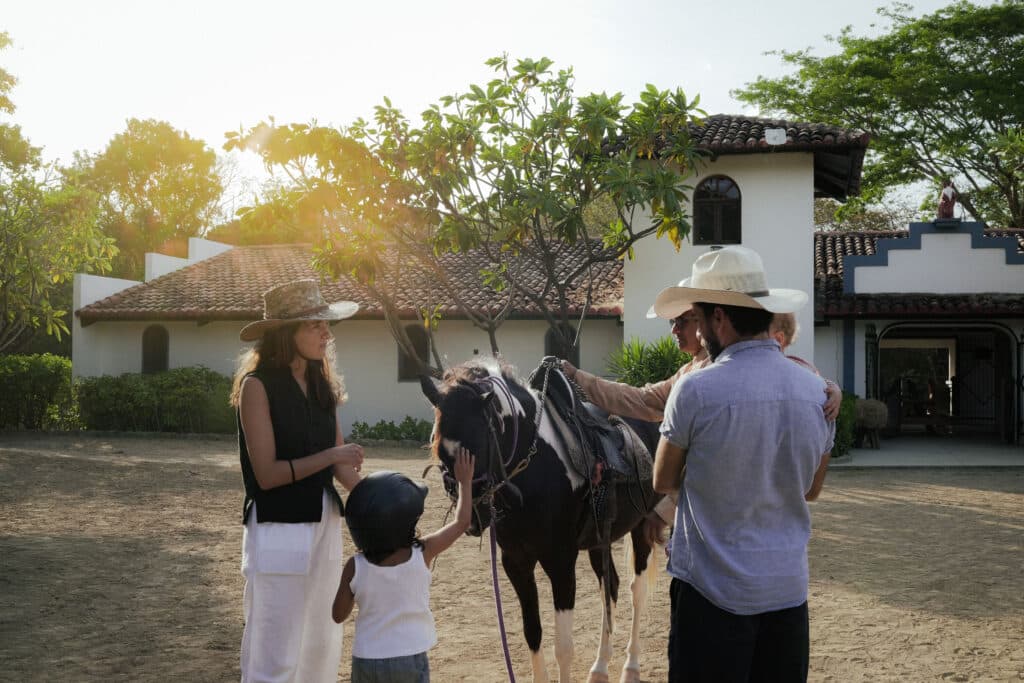 A group of adults and a child stand around a saddled horse in front of a house with a tiled roof. On this Family Trip Nicaragua adventure, the child, wearing a helmet, holds the horse’s reins as sunlight filters through the trees.