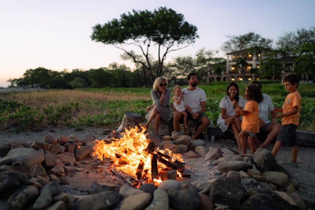 A group of adults and children sit on logs around a campfire on a beach at sunset, with trees and a house visible in the background.