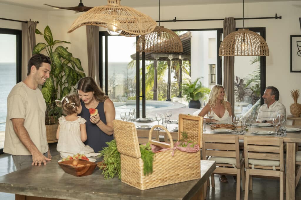 A family gathers indoors near a kitchen island with fruit and a picnic basket, while another couple sits at a dining table by large windows overlooking a pool and the ocean.