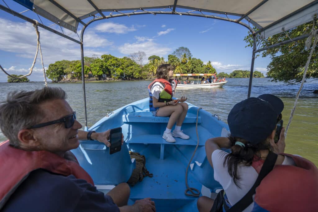 Three people wearing life jackets sit in a small blue boat on a lake, capturing photos of another boat and a lush green island under a bright blue sky with scattered clouds—an ideal scene for memorable multi-generational family vacations.
