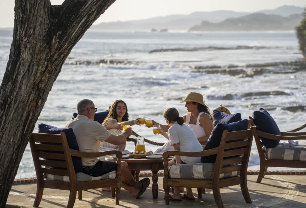 Five people sit on outdoor sofas by the ocean at Rancho Santana, toasting with drinks around a low table filled with food. The scene is sunny and relaxed, capturing the laid-back spirit of Nicaragua’s premier anti-resort, with waves and distant hills in the background.