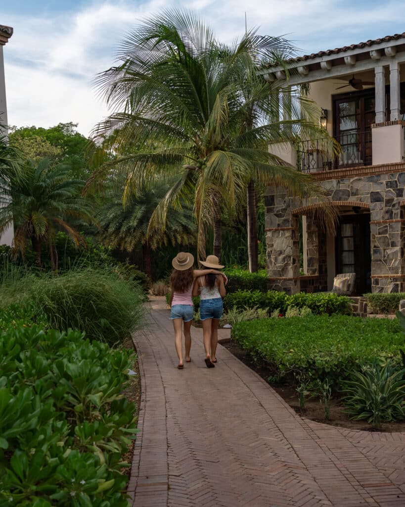 Two girls in shorts and sun hats walk down a brick path surrounded by lush greenery and palm trees, passing by a stone building with a balcony on a sunny day—a perfect setting to soak up the sunshine and enjoy every moment with this limited time offer.