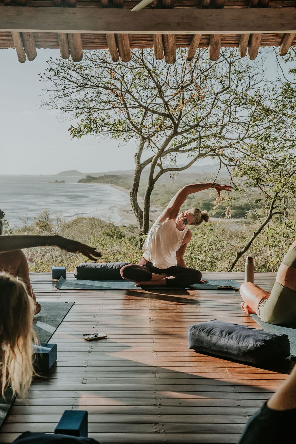 At a FloYo Retreat, a yoga instructor leads a seated side stretch on a wooden deck overlooking a scenic beach and ocean, surrounded by trees and students on yoga mats as warm sunlight fills the open-air space—Trevor Hall playing softly in the background.